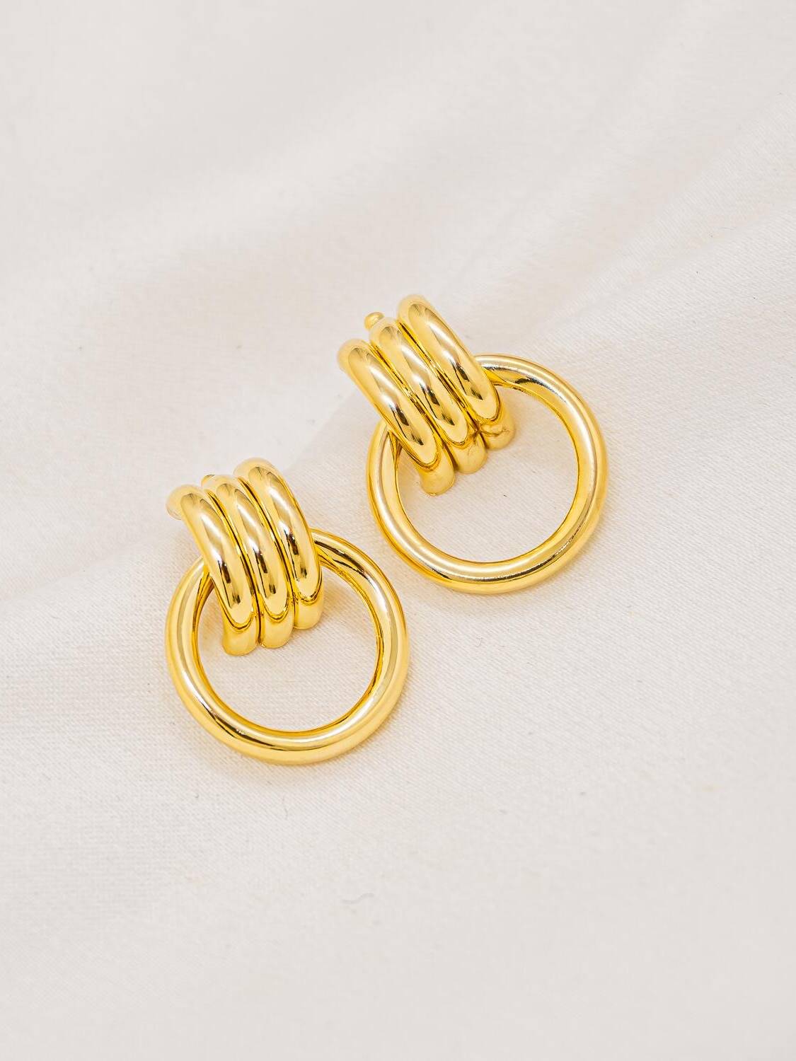Boucles d’oreilles dorées lisses, base en bandes arrondies et grand anneau ajouré, sur tissu clair
