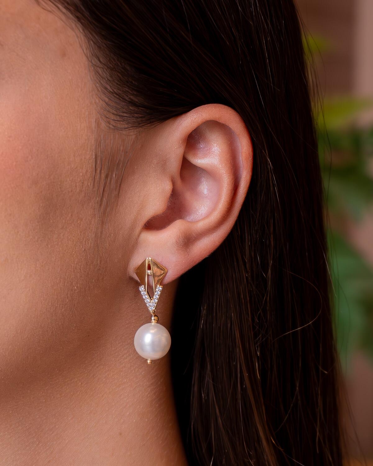 Boucle d’oreille dorée à losange ajouré, V serti de pierres claires et perle ronde pendante.