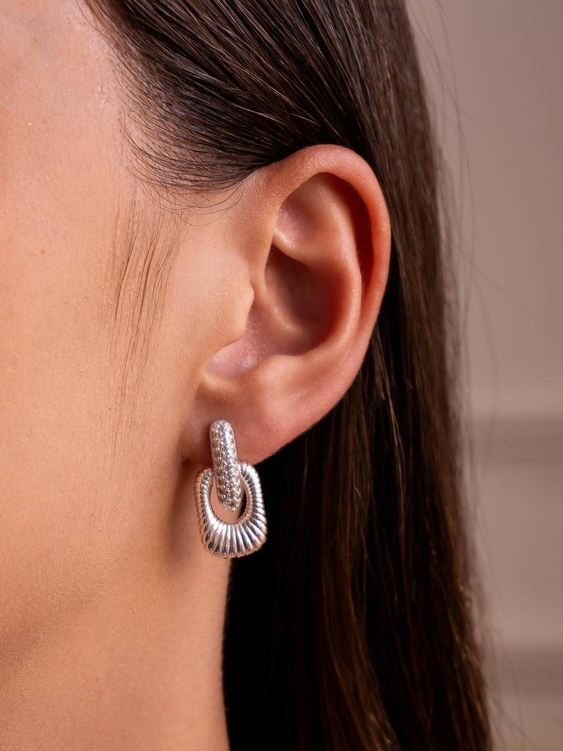 Boucle d’oreille argentée portée : barrette sertie de pierres claires et rectangle ajouré strié suspendu.