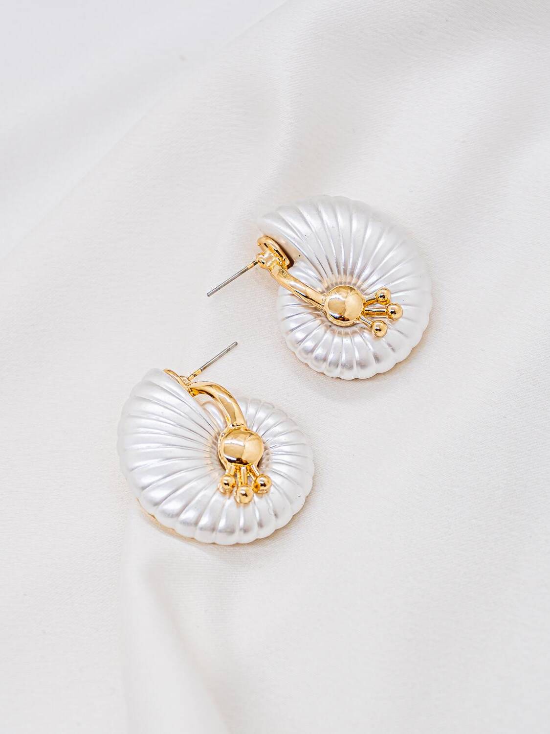 Paire de boucles d’oreilles coquille blanc irisé strié, cœur doré à petites billes, sur tissu clair.