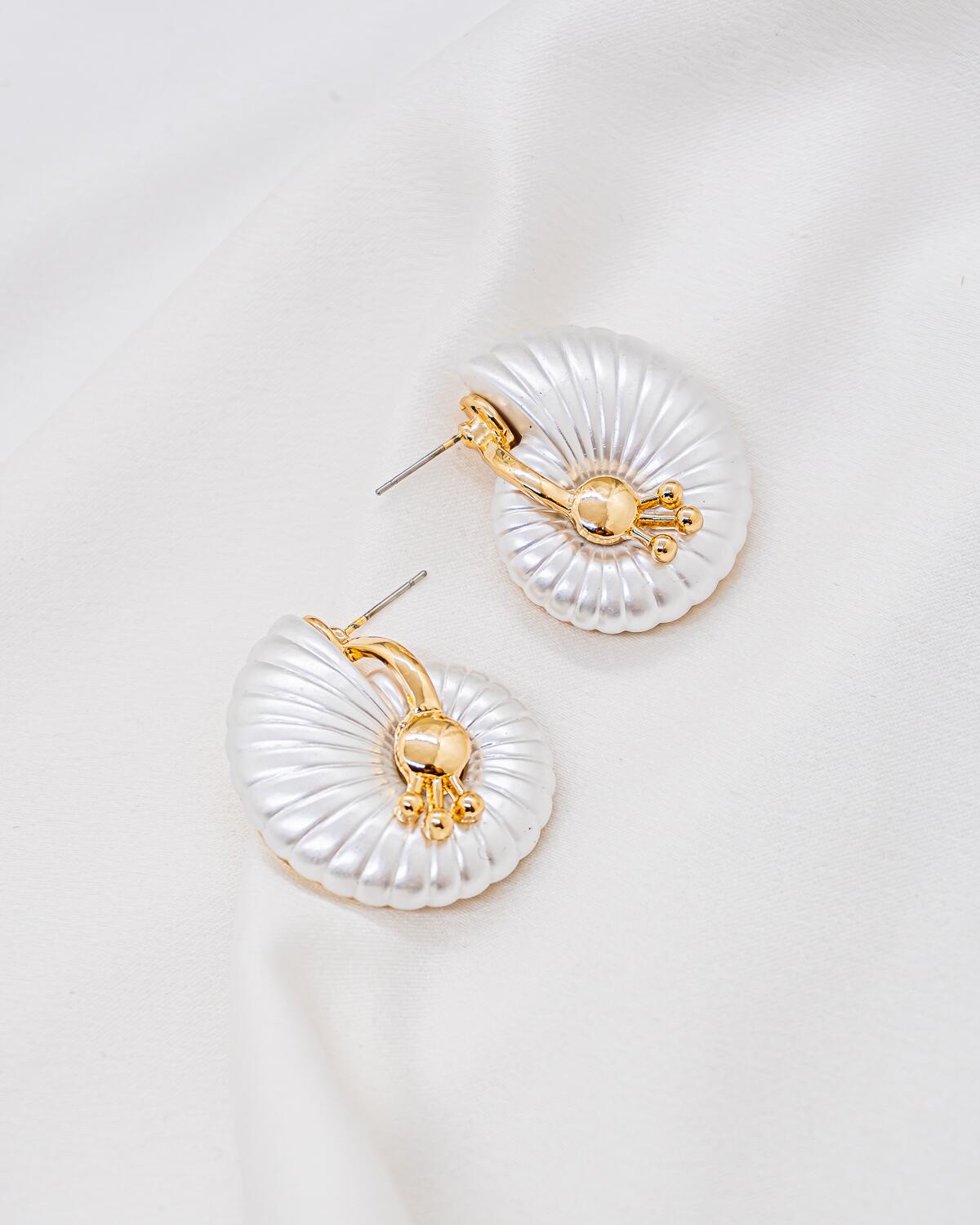 Paire de boucles d’oreilles coquille blanc irisé strié, cœur doré à petites billes, sur tissu clair.