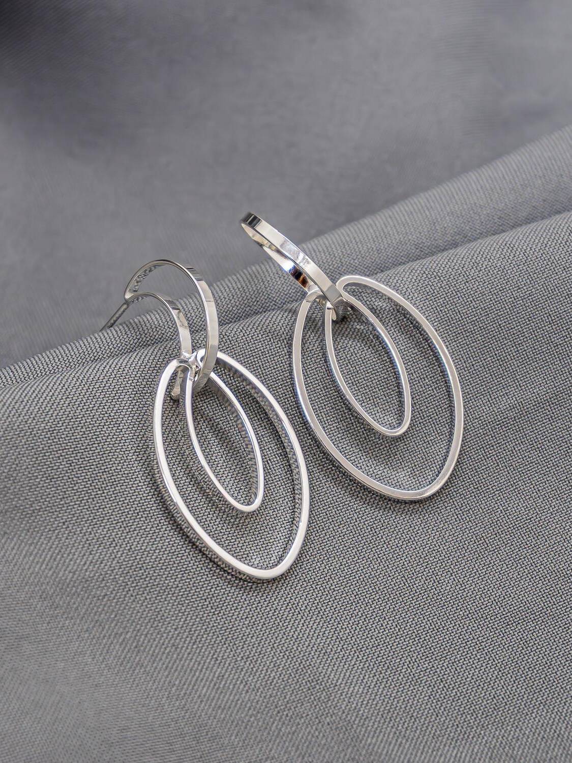 Boucles d’oreilles argentées, demi-anneau plat et deux ovales ajourés emboîtés, sur tissu gris