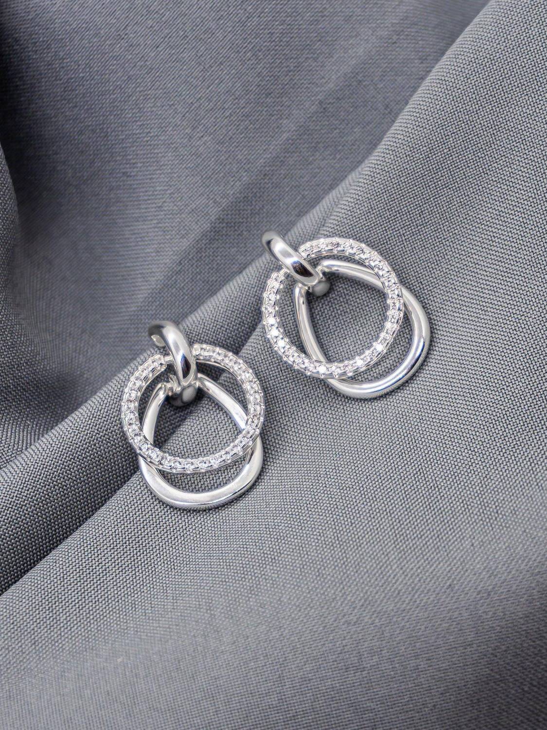 Boucles d’oreilles argentées, anneau serti de pierres claires croisé avec un anneau lisse, sur tissu gris