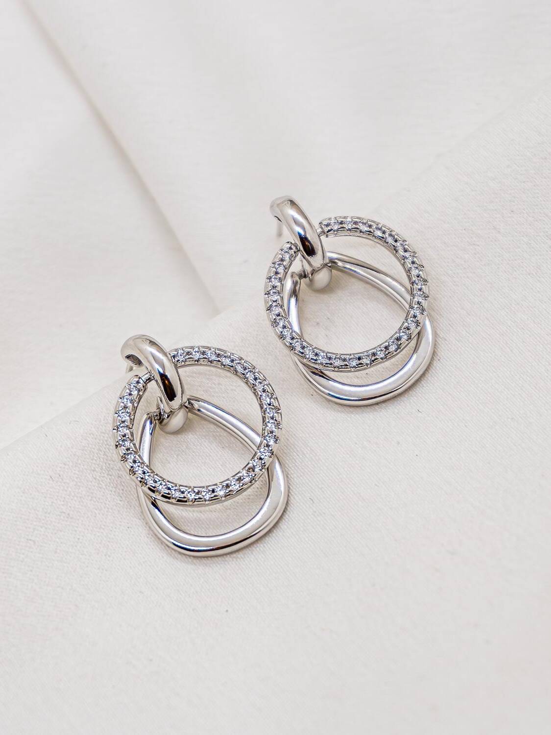 Boucles d’oreilles argentées, anneau serti de pierres claires croisant un anneau lisse, sur tissu crème