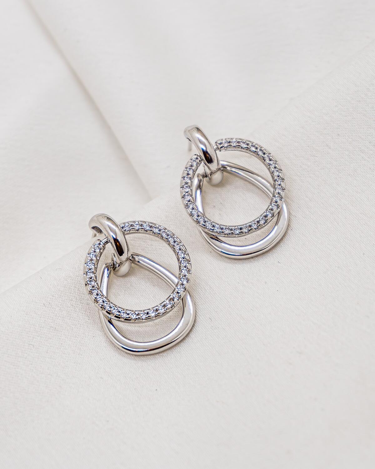Boucles d’oreilles argentées, anneau serti de pierres claires croisant un anneau lisse, sur tissu crème