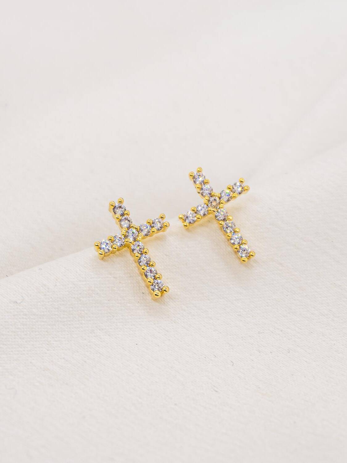 Paire de petites boucles d’oreilles croix dorées serties de pierres claires, sur tissu blanc