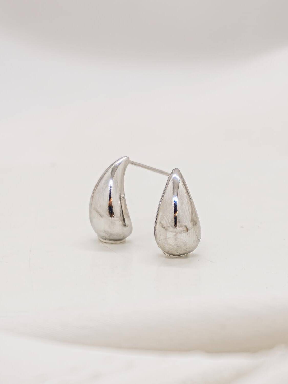 Paire de très petites boucles d’oreilles argentées en goutte lisse, au reflet brillant sur fond ivoire.
