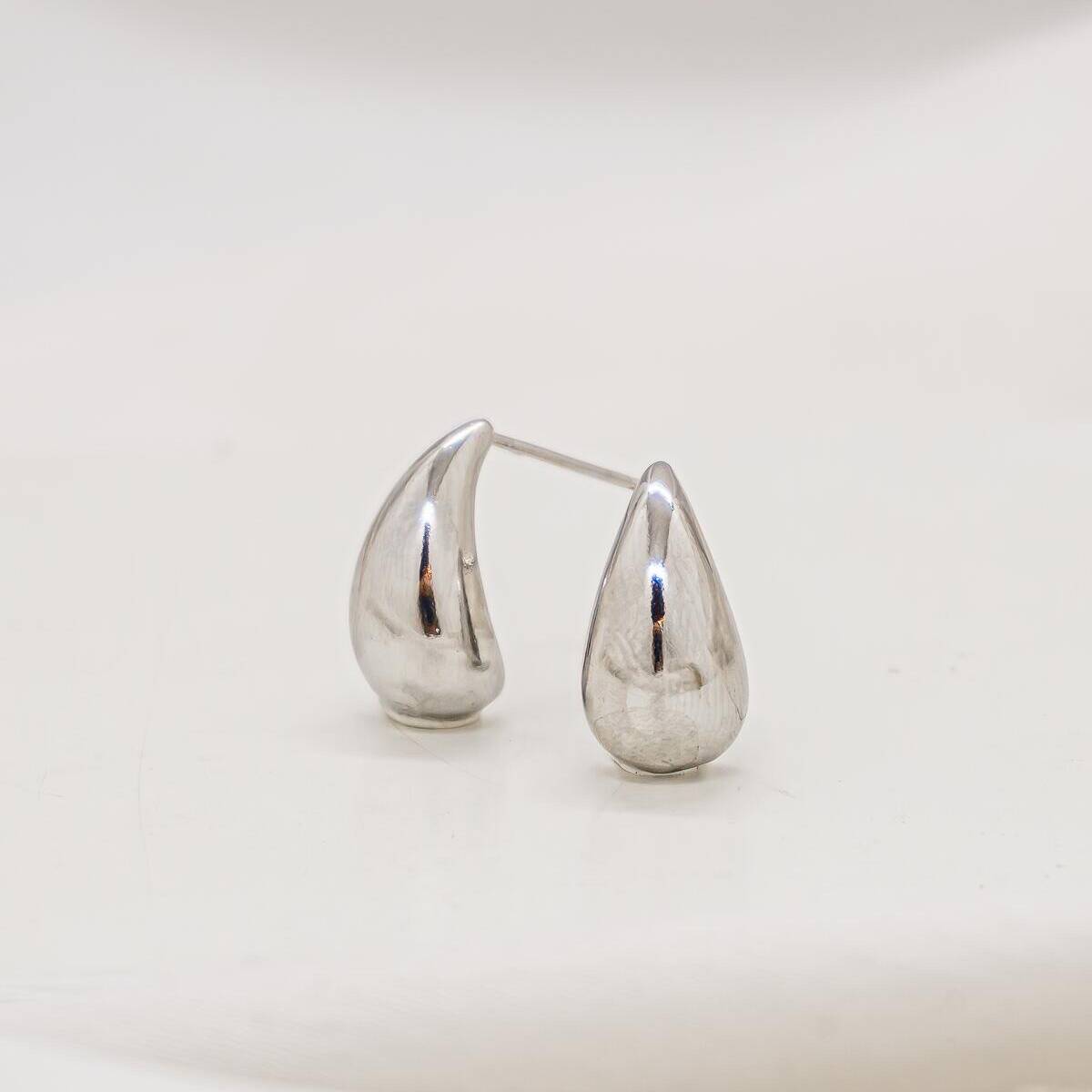 Pair of tiny smooth silver-toned teardrop earrings, with a glossy shine against an ivory background.