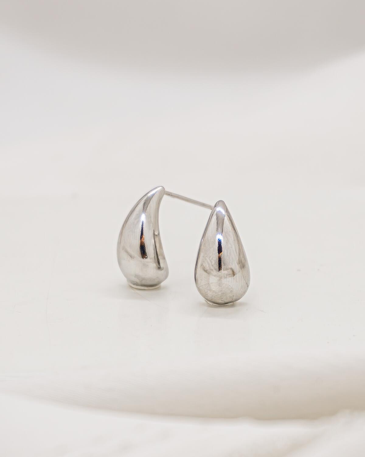 Paire de très petites boucles d’oreilles argentées en goutte lisse, au reflet brillant sur fond ivoire.