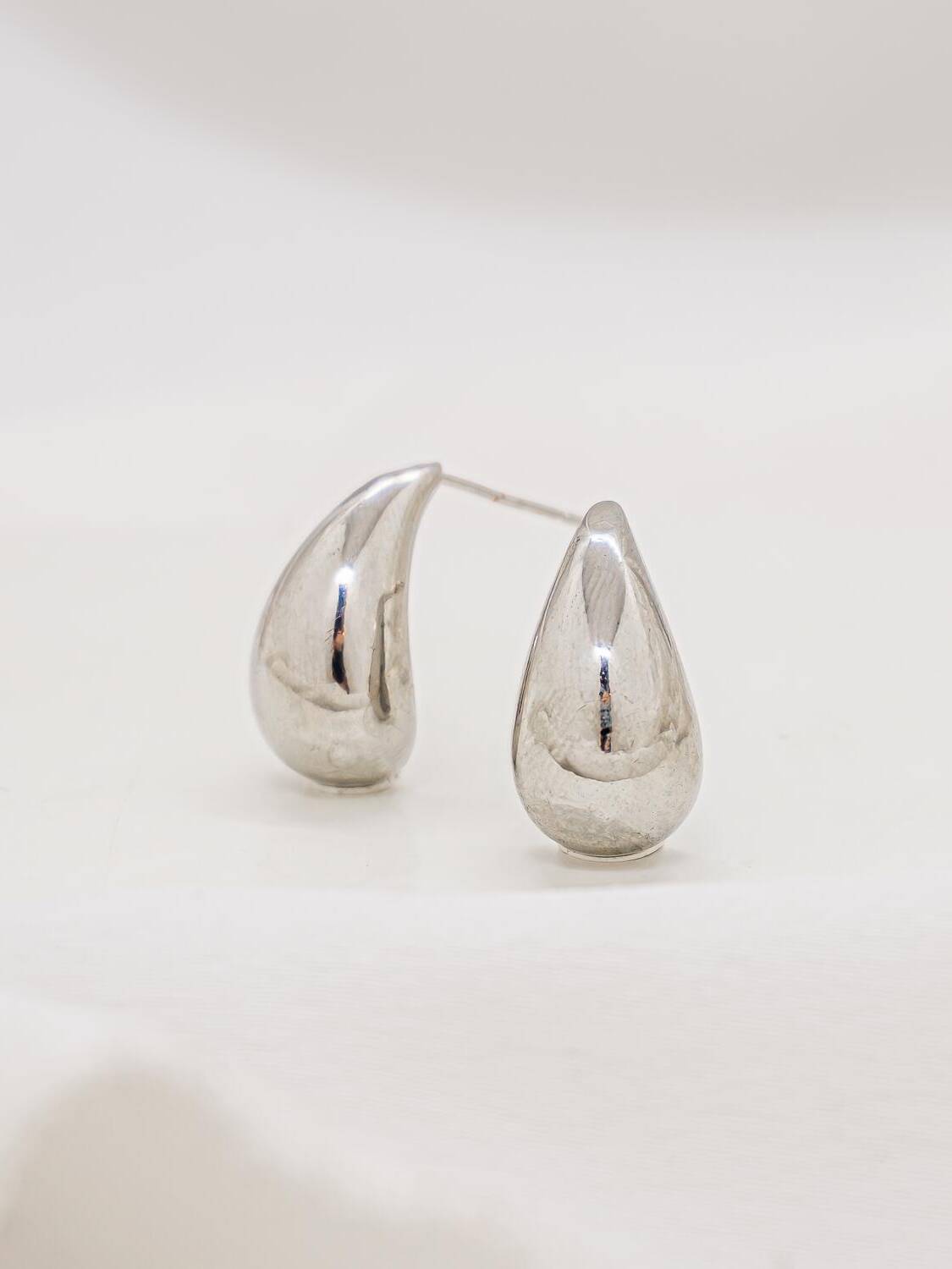 Paire de boucles d’oreilles argentées compactes en forme de goutte lisse, posées sur un tissu clair.