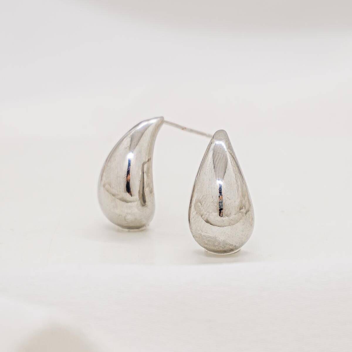 Compact silver-toned smooth teardrop earrings pair, placed on a light fabric.