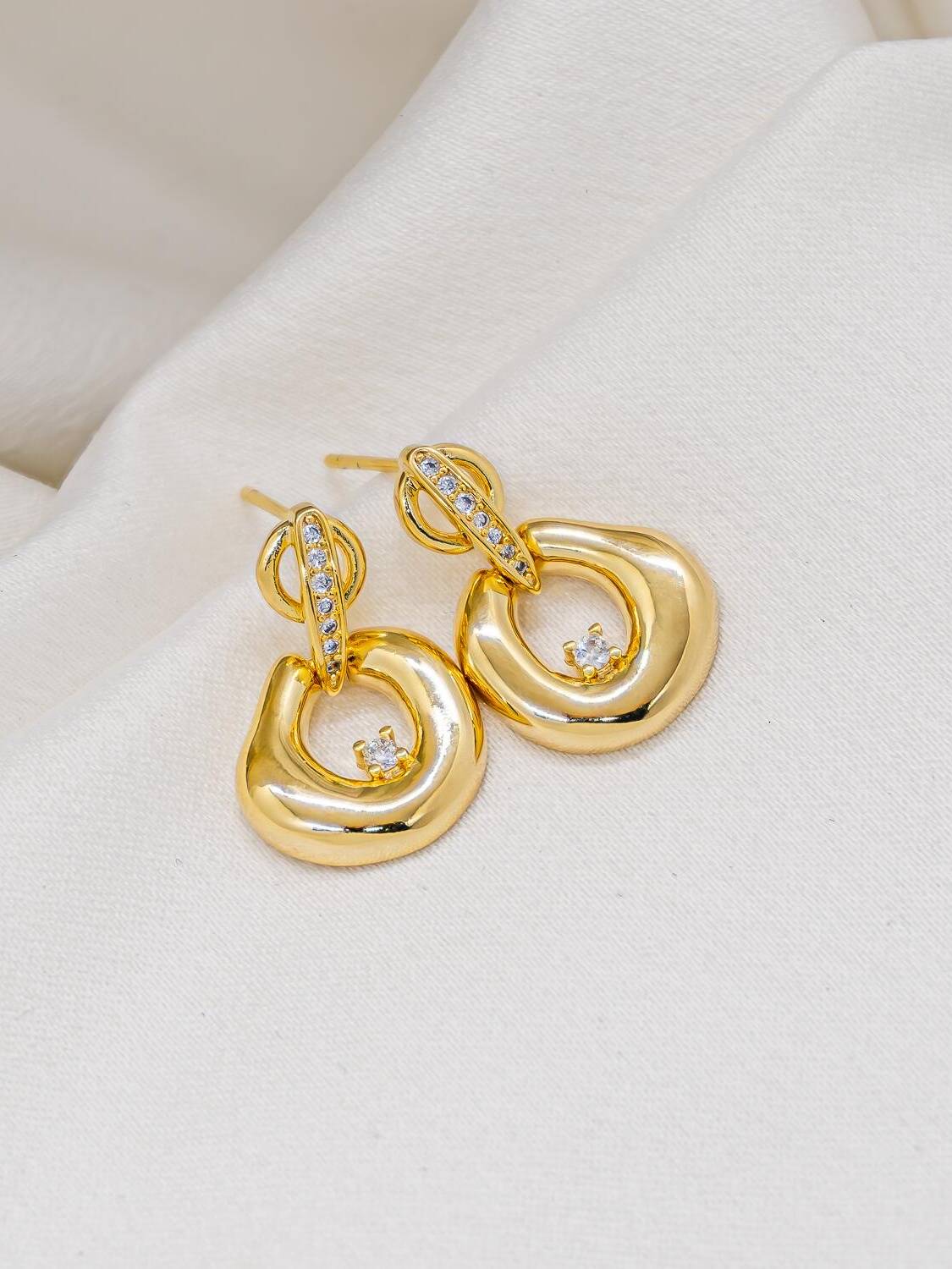 Boucles d’oreilles dorées à pendentif organique ajouré, pierre claire et barrette sertie, sur tissu crème