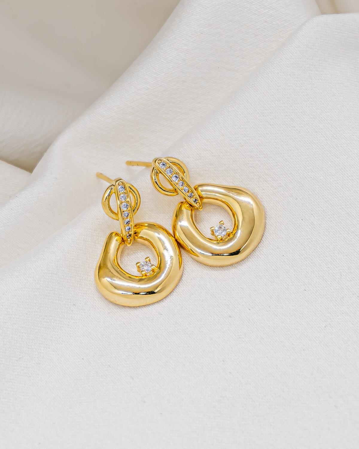 Boucles d’oreilles dorées à pendentif organique ajouré, pierre claire et barrette sertie, sur tissu crème