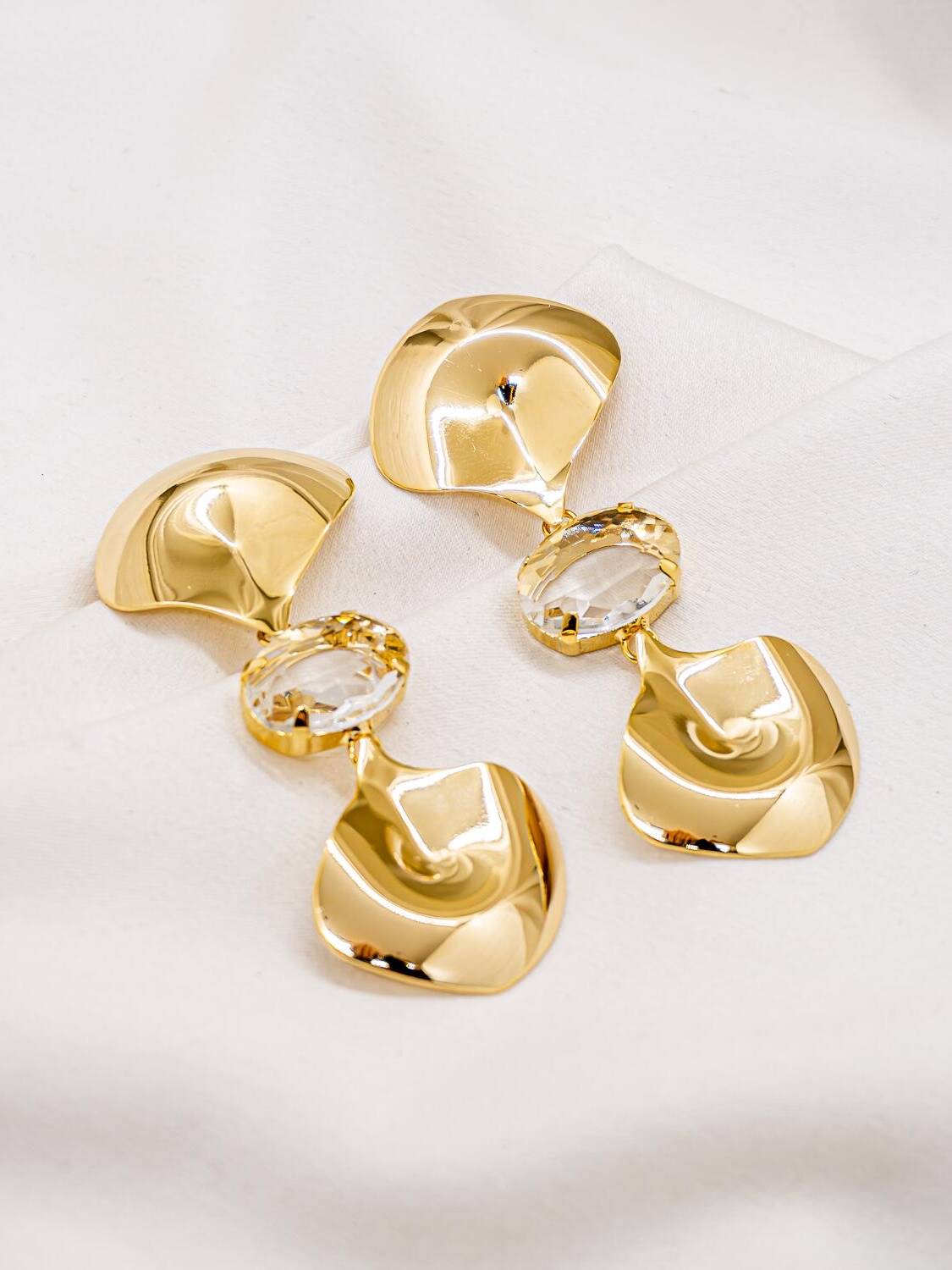Paire de boucles d’oreilles dorées aux plaques bombées organiques, séparées par un grand cristal ovale clair.