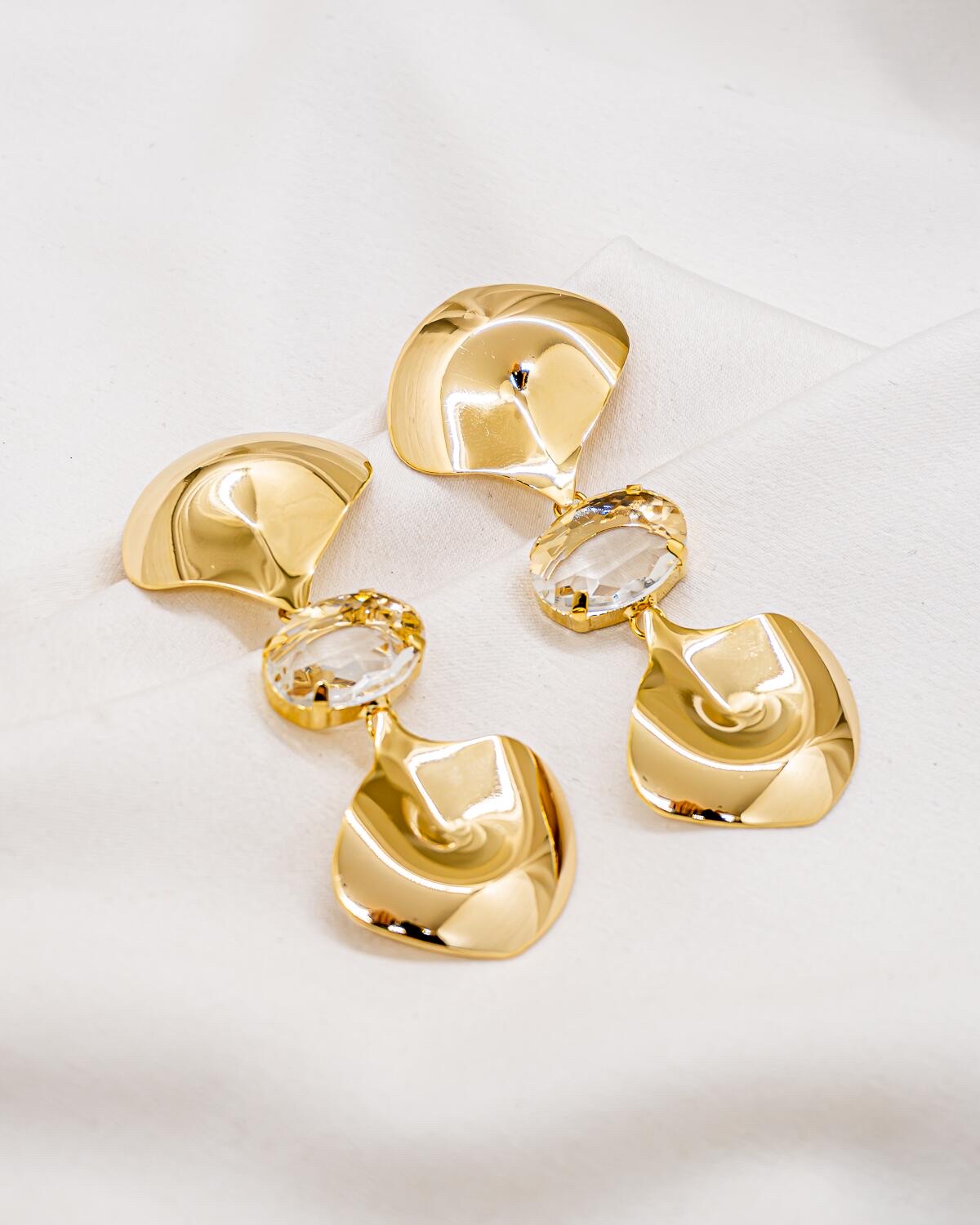 Paire de boucles d’oreilles dorées aux plaques bombées organiques, séparées par un grand cristal ovale clair.