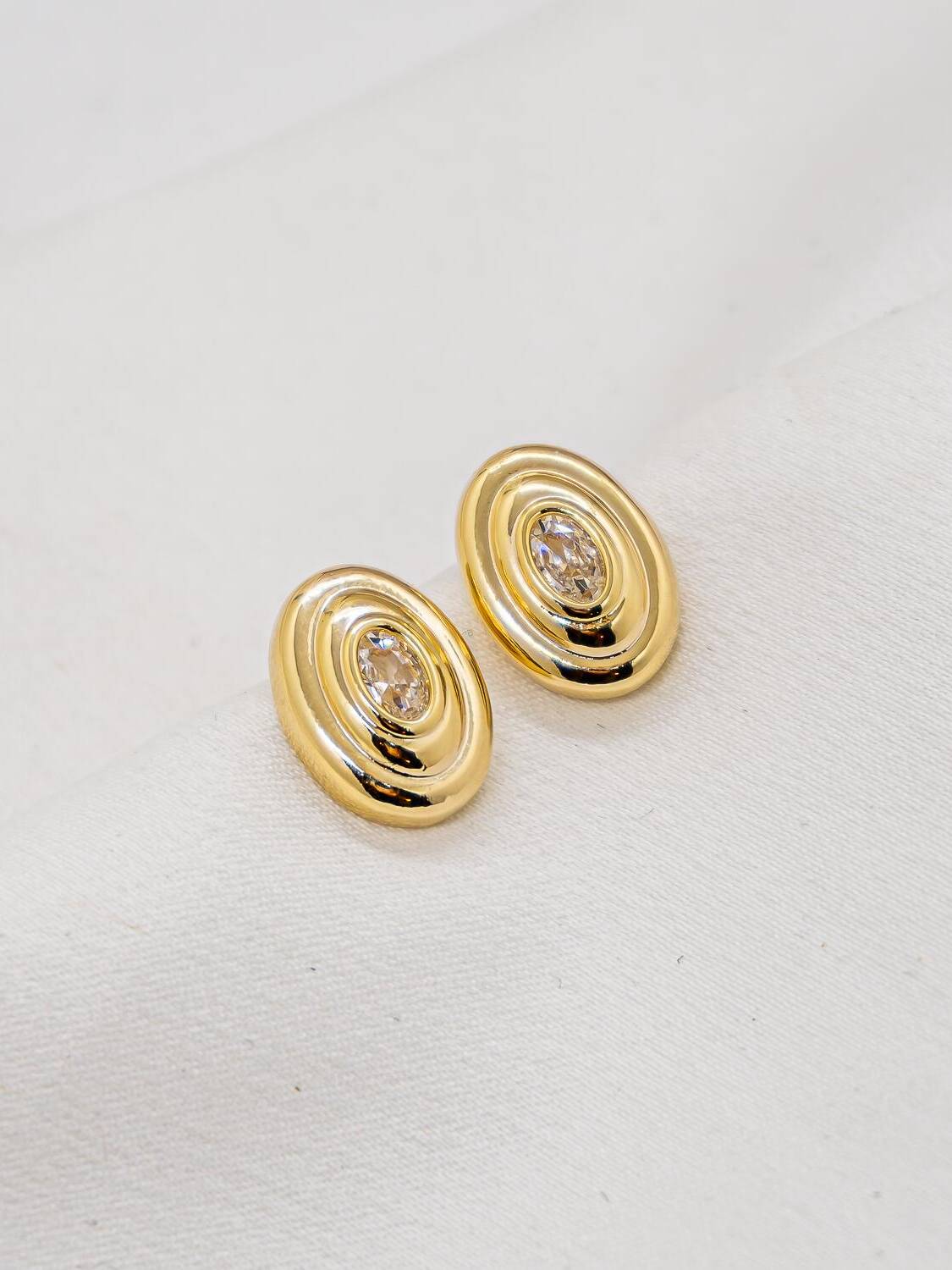 Paire de boucles d’oreilles ovales dorées à reliefs concentriques et pierre claire centrale