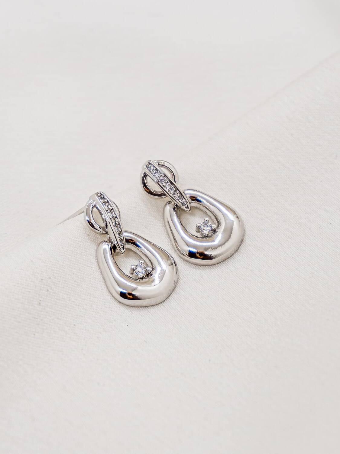 Boucles d’oreilles argentées, pendentif souple ajouré avec pierre claire et barrette sertie, sur tissu crème