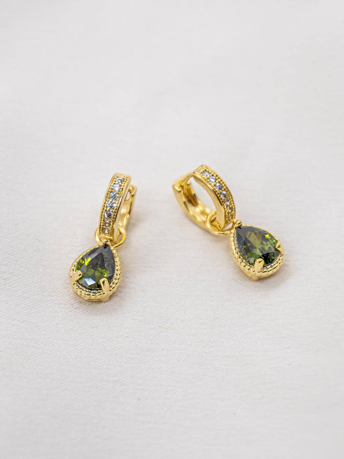 Petites créoles dorées serties de pierres claires, avec une goutte verte pendante, sur tissu clair