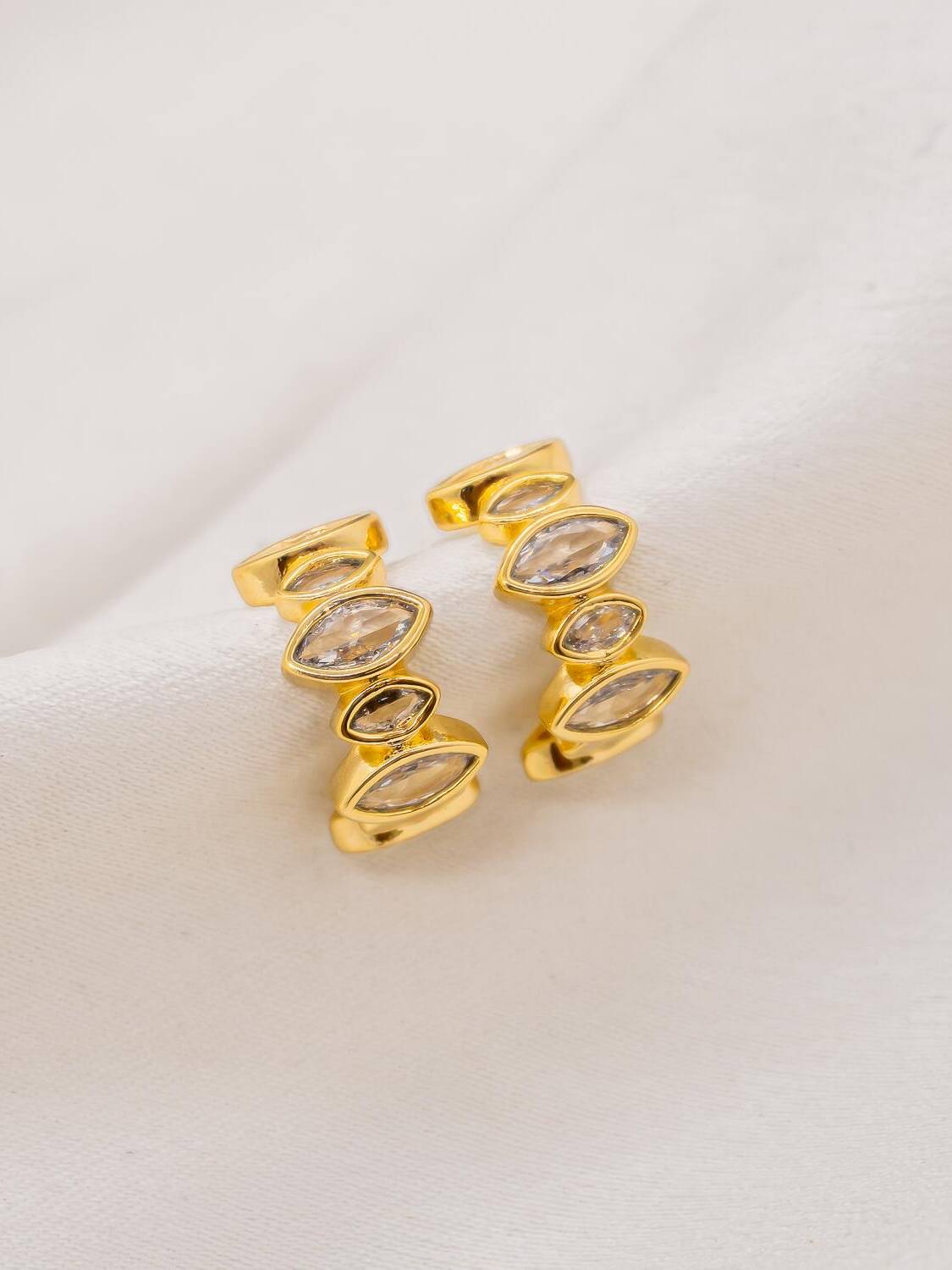 Paire de petites créoles dorées ornées de pierres claires en navette, sur tissu satiné ivoire.