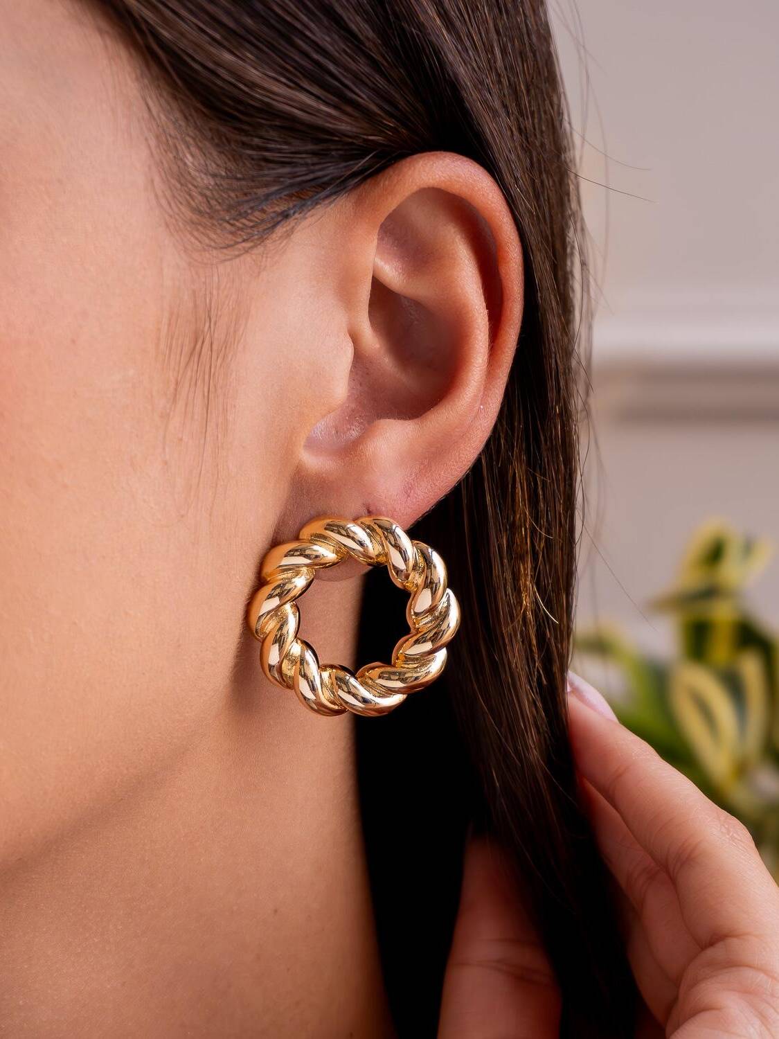 Boucle d’oreille dorée en anneau ajouré aux brins torsadés, portée sur fond de feuillage.