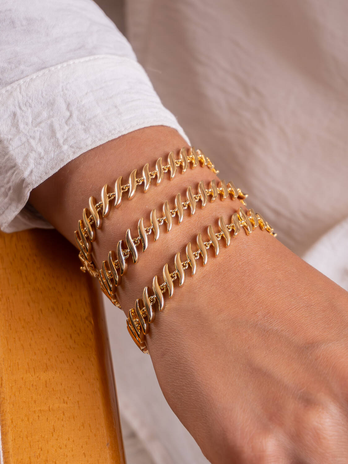 Trois bracelets dorés à maillons allongés et incurvés, superposés sur un poignet, devant une chemise claire