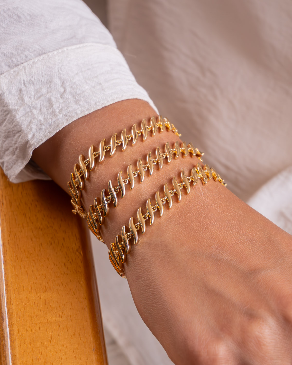Trois bracelets dorés à maillons allongés et incurvés, superposés sur un poignet, devant une chemise claire