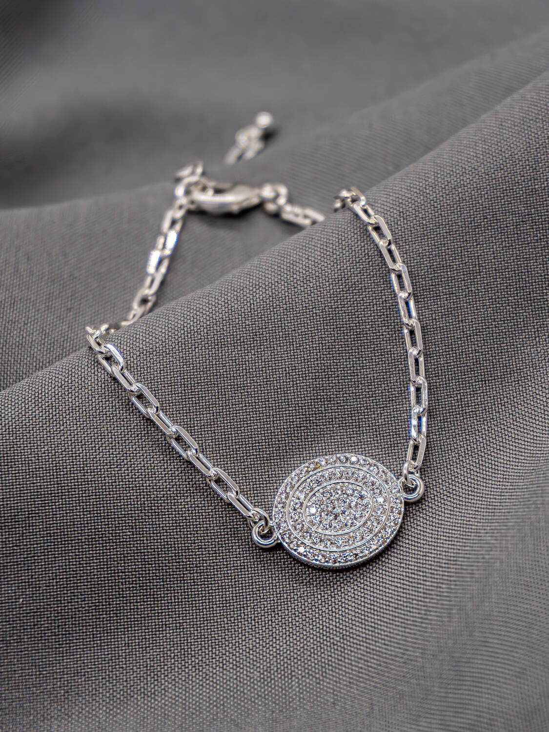 Bracelet argenté à maillons rectangulaires avec pendentif ovale pavé de pierres claires, sur tissu gris