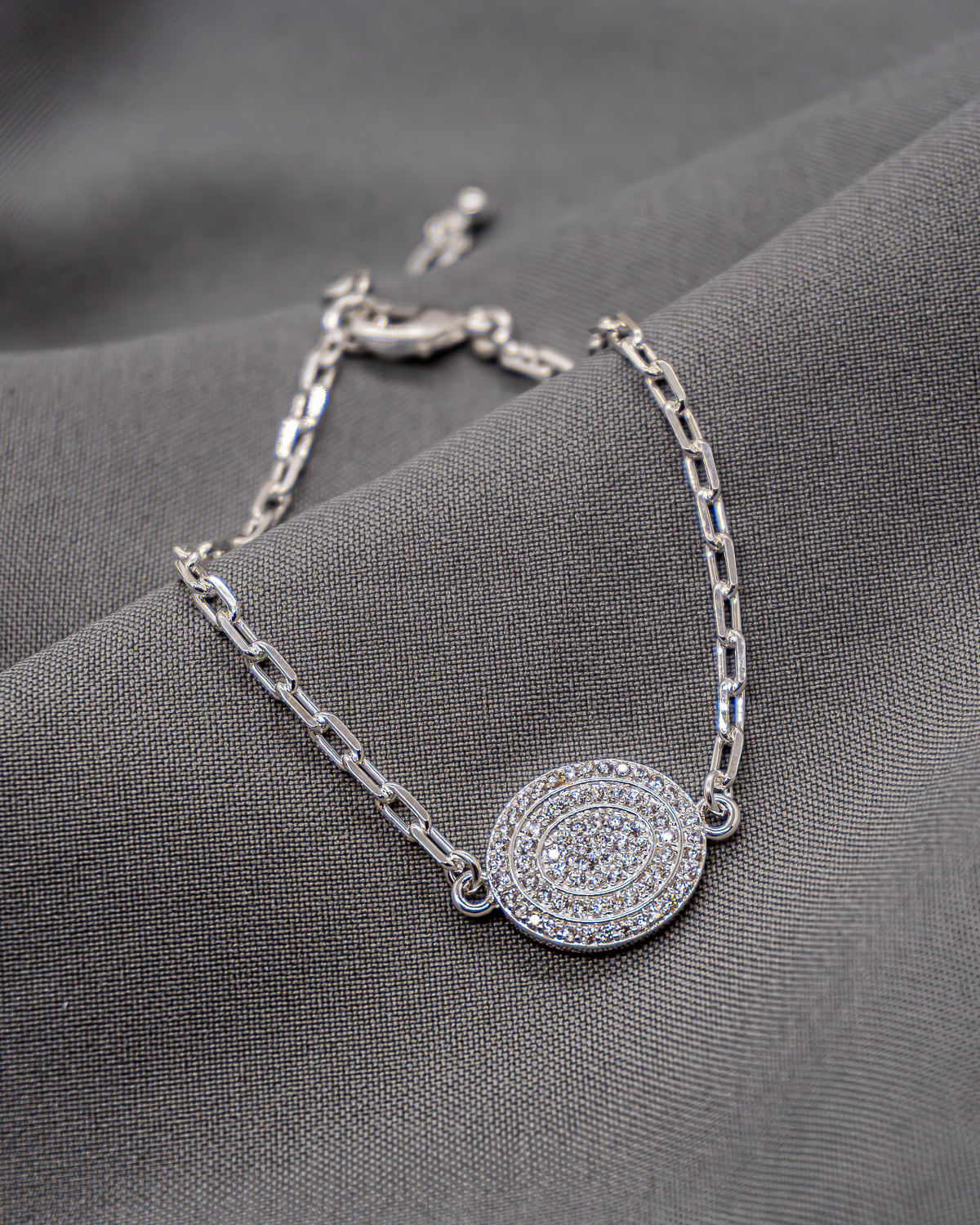 Pulseira prateada de elos retangulares com pingente oval cravejado de pedras claras, sobre tecido cinza