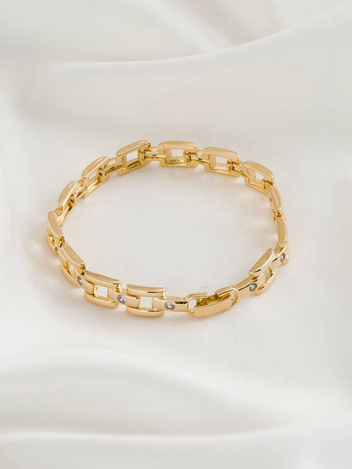 Bracelet doré à maillons rectangulaires ajourés avec quelques pierres claires serties, posé sur un tissu crème
