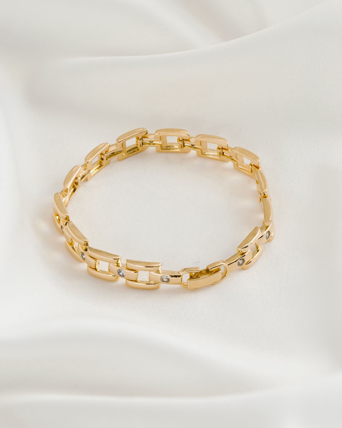 Bracelet doré à maillons rectangulaires ajourés avec quelques pierres claires serties, posé sur un tissu crème