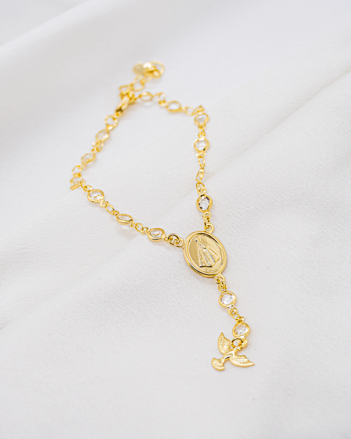 Pulseira dourada com pedras claras cravejadas, medalha oval Nossa Senhora e pequena pomba pendente