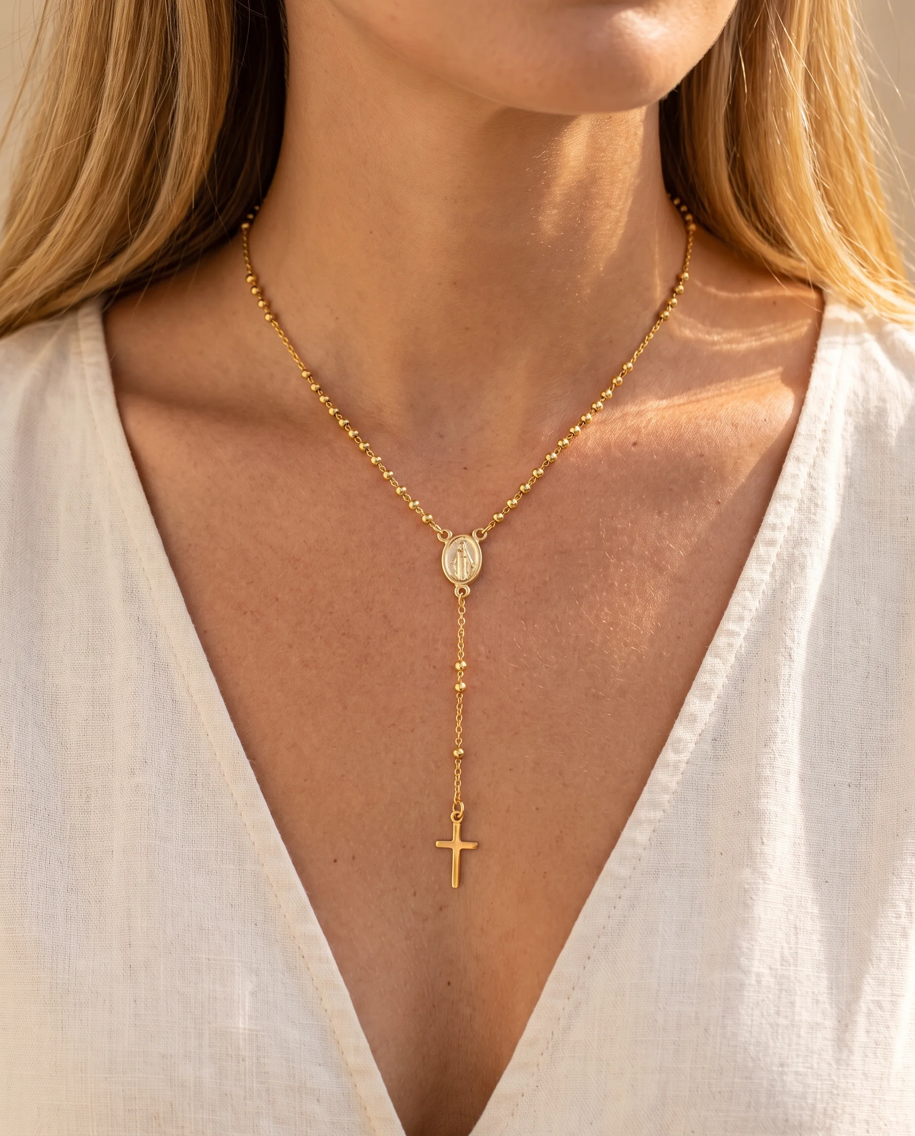 Gold rosary-style necklace worn in a Y-shape, medal at the hollow of the neckline and cross as a pendant