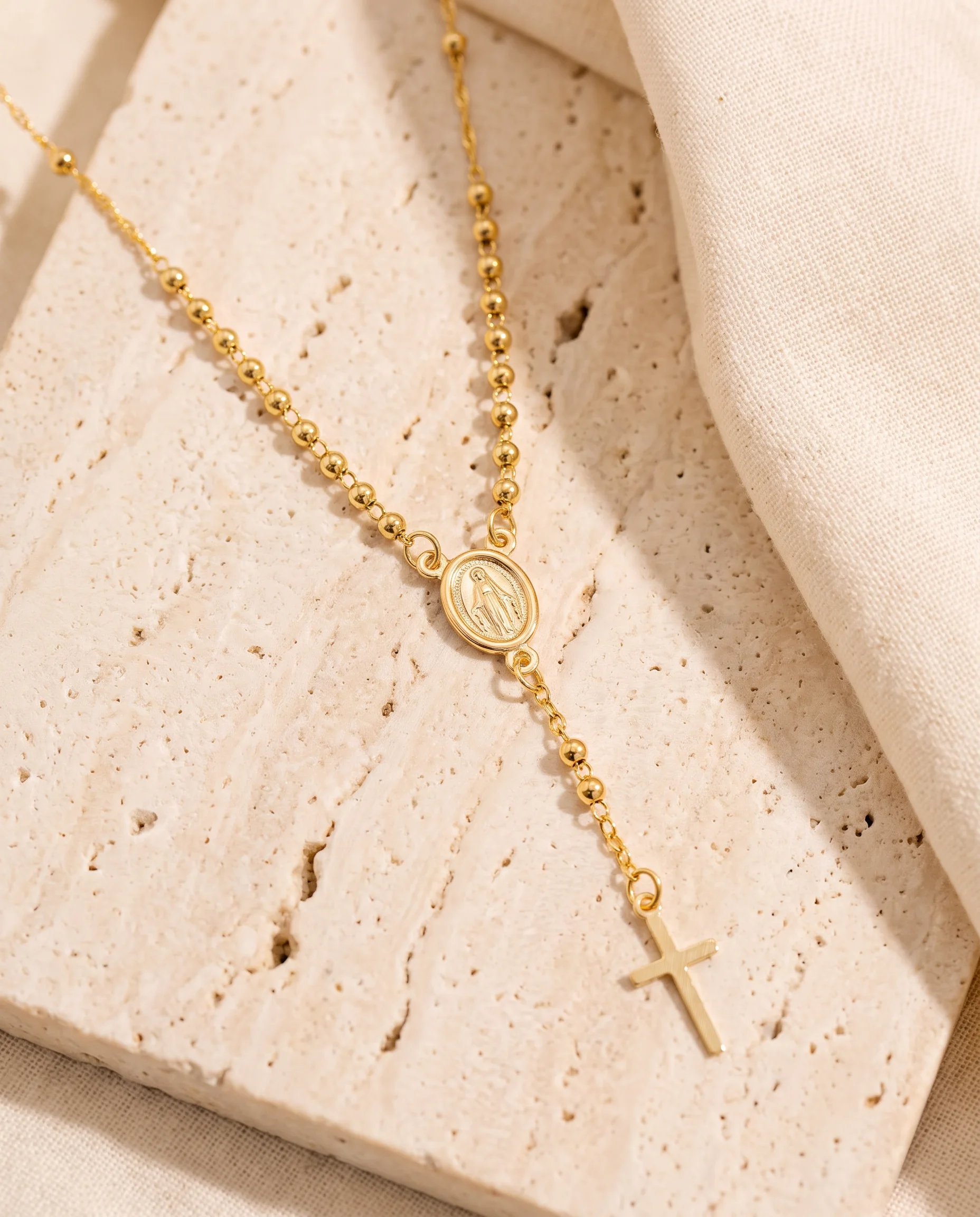 Gold rosary-style necklace with oval medal and small cross, laid on travertine