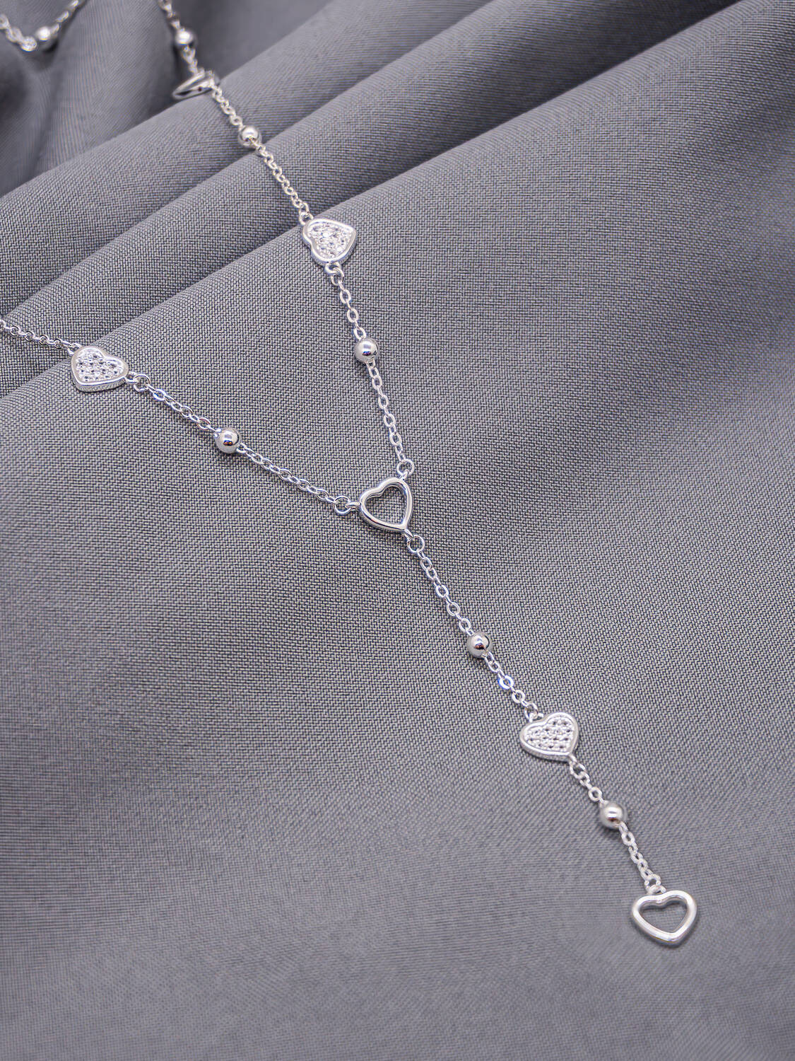 Collier argenté forme cravate à cœurs lisses et cœurs sertis, posé sur un tissu gris