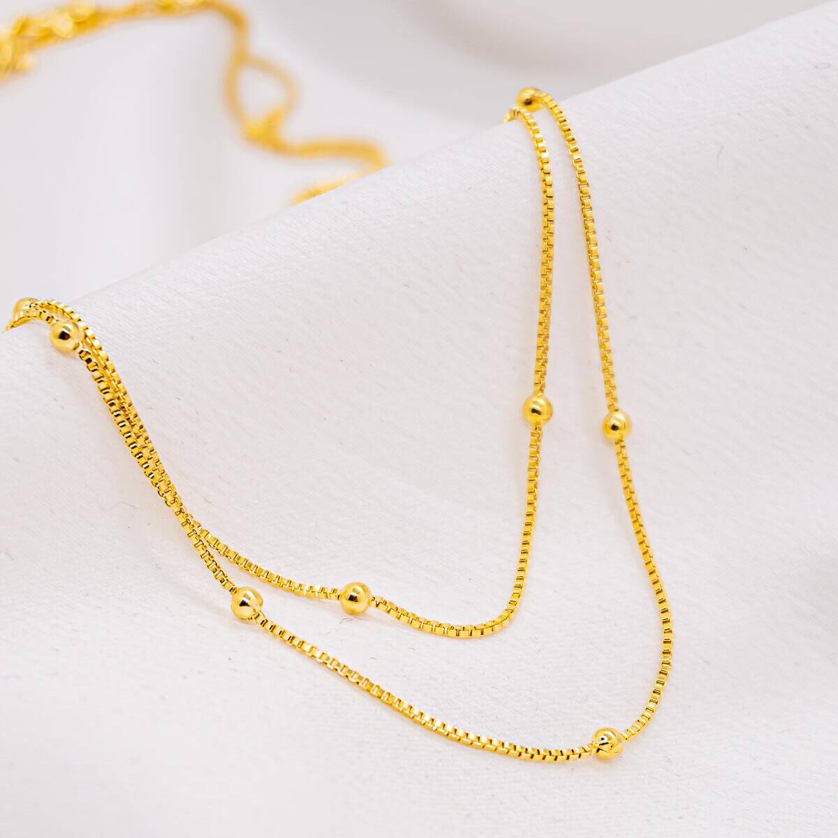 Long gold necklace in fine doubled Venetian chain, with small smooth beads spaced apart, on white fabric