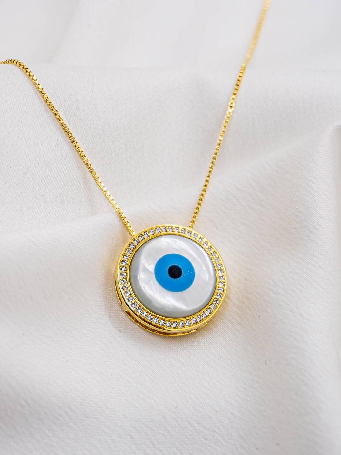 Pendentif rond en nacre avec œil grec bleu et anneau de pierres claires, sur chaîne dorée