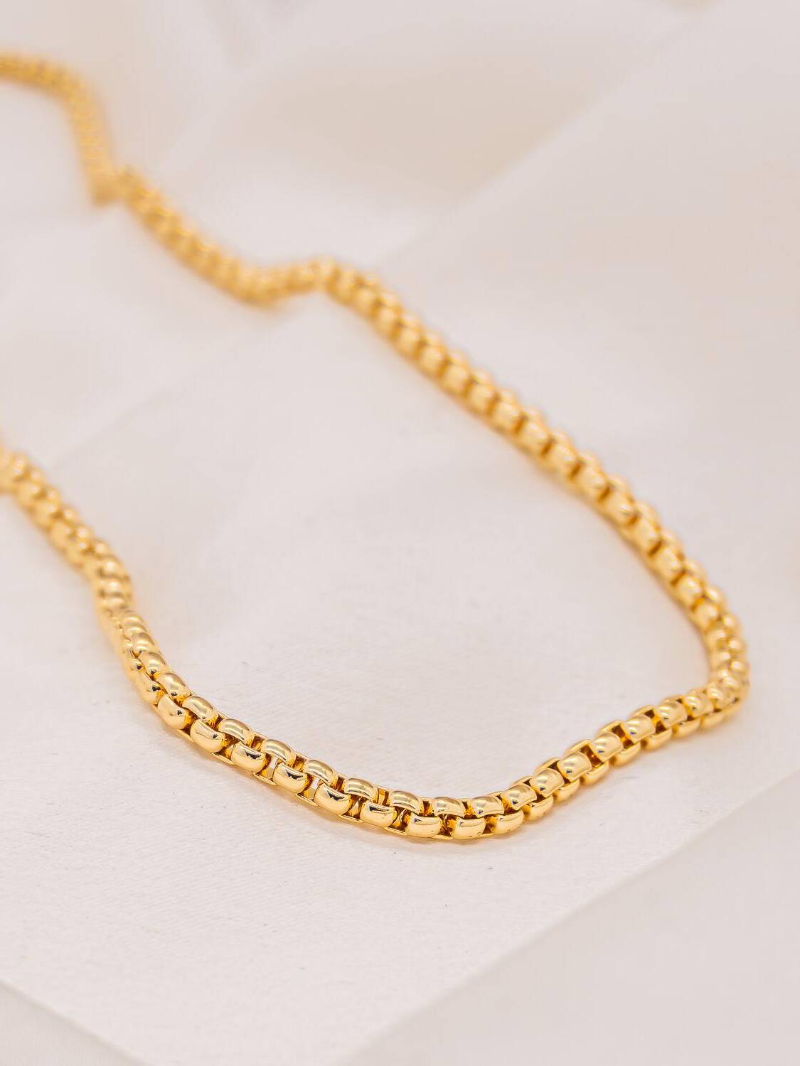 Collier doré en maillons carrés entrelacés et polis, posé en courbe sur un tissu clair