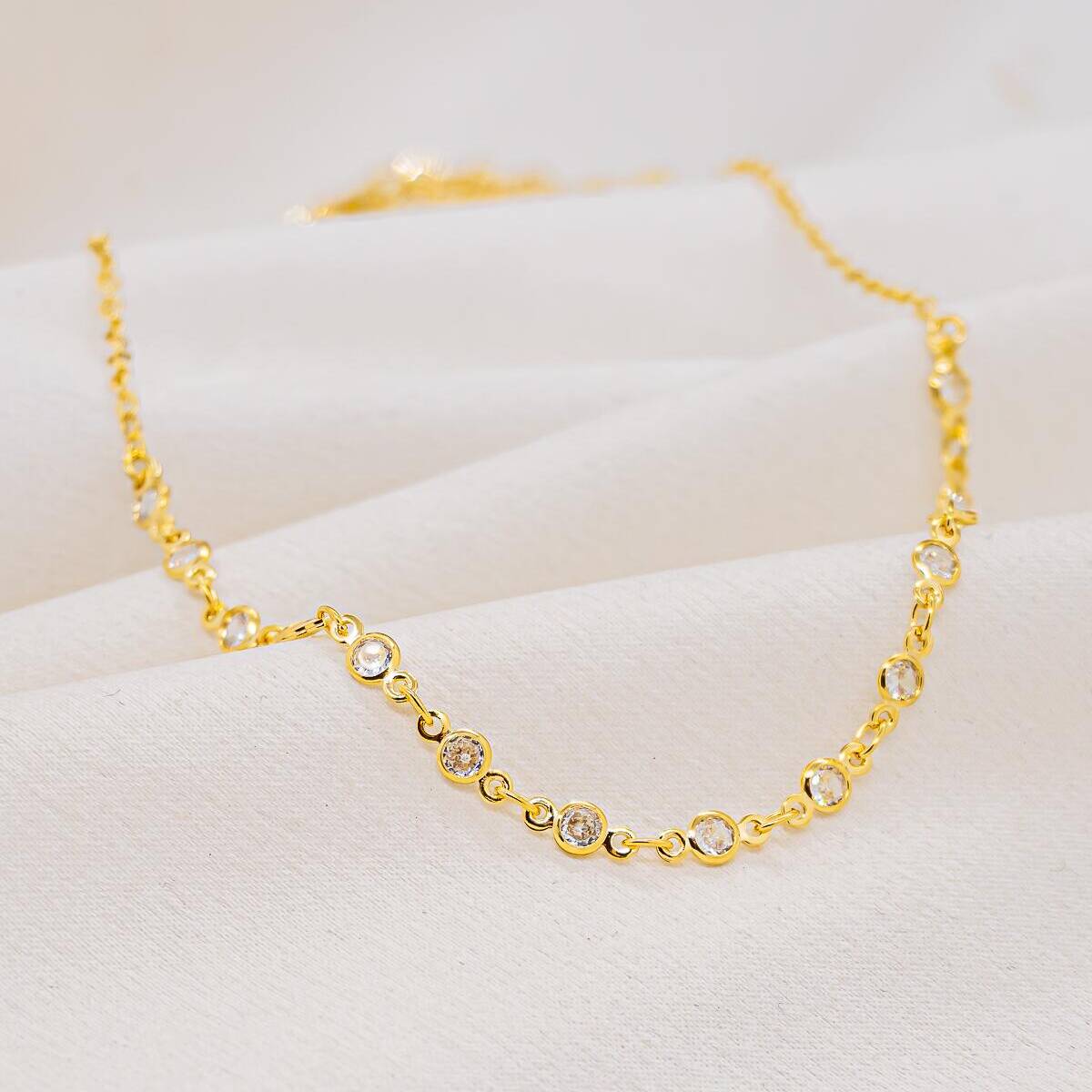 Gold choker made of small round links, each set with a clear stone, on white fabric