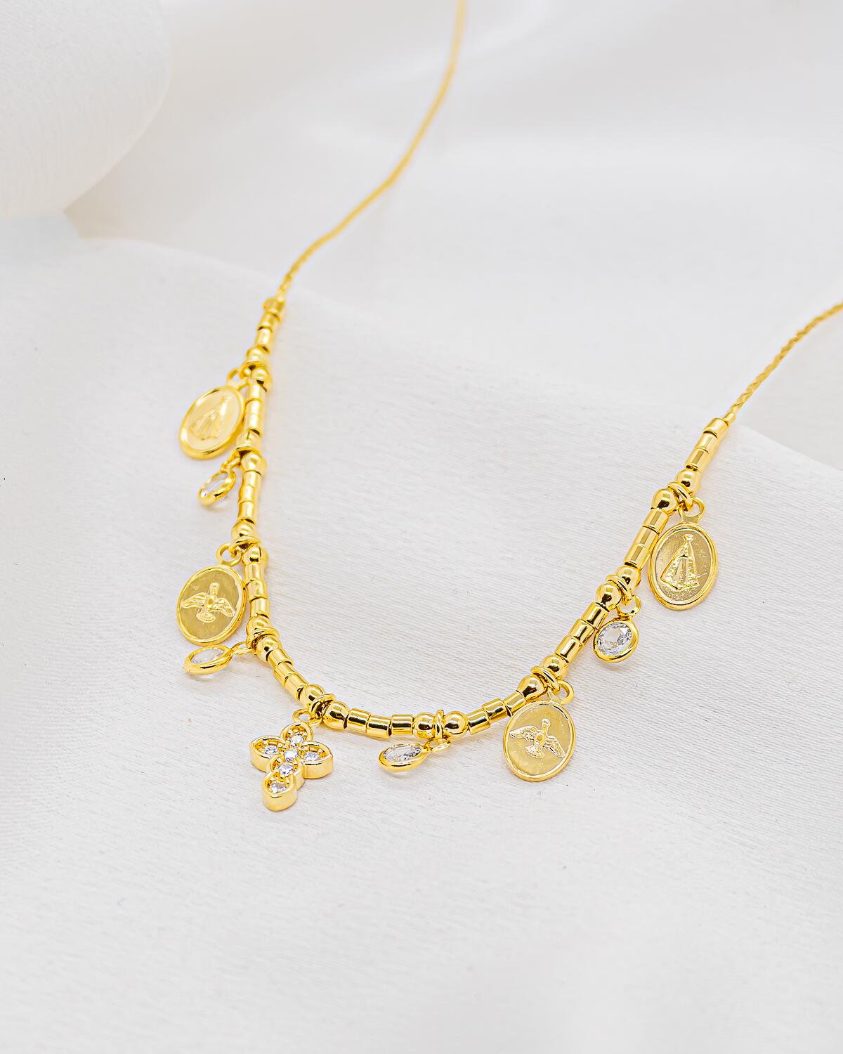 Colar gargantilha dourado com pequenos tubos, medalhas religiosas ovais, pedras claras pendentes e cruz cravejada