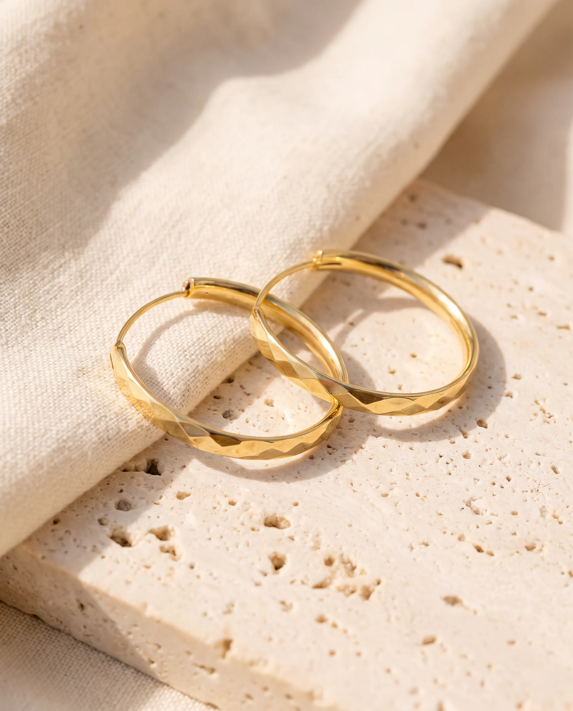 Pair of gold faceted hoop earrings with diamond-shaped facets placed on a light stone and ecru linen