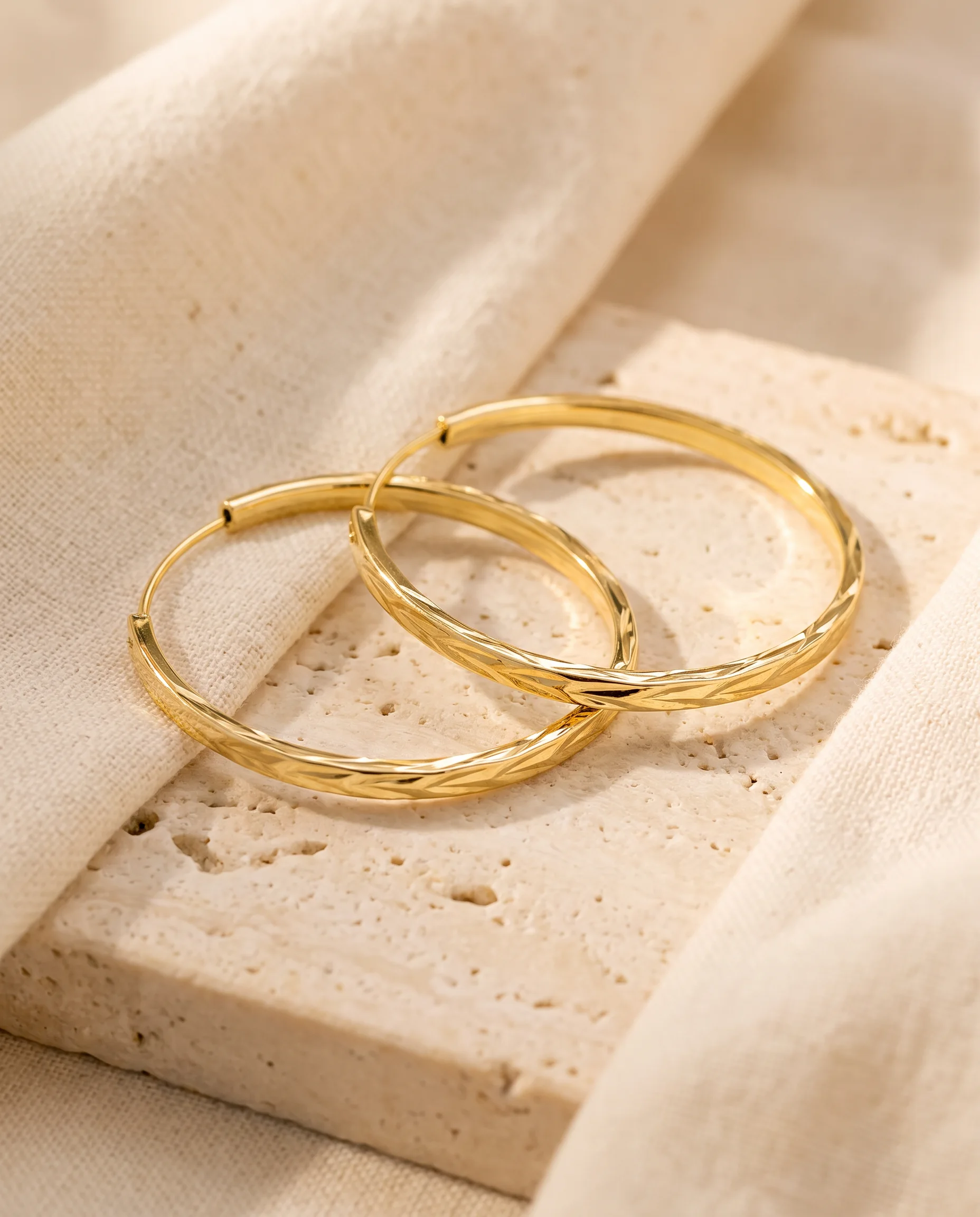 Fine gold hoop earrings engraved with chevrons, laid on travertine