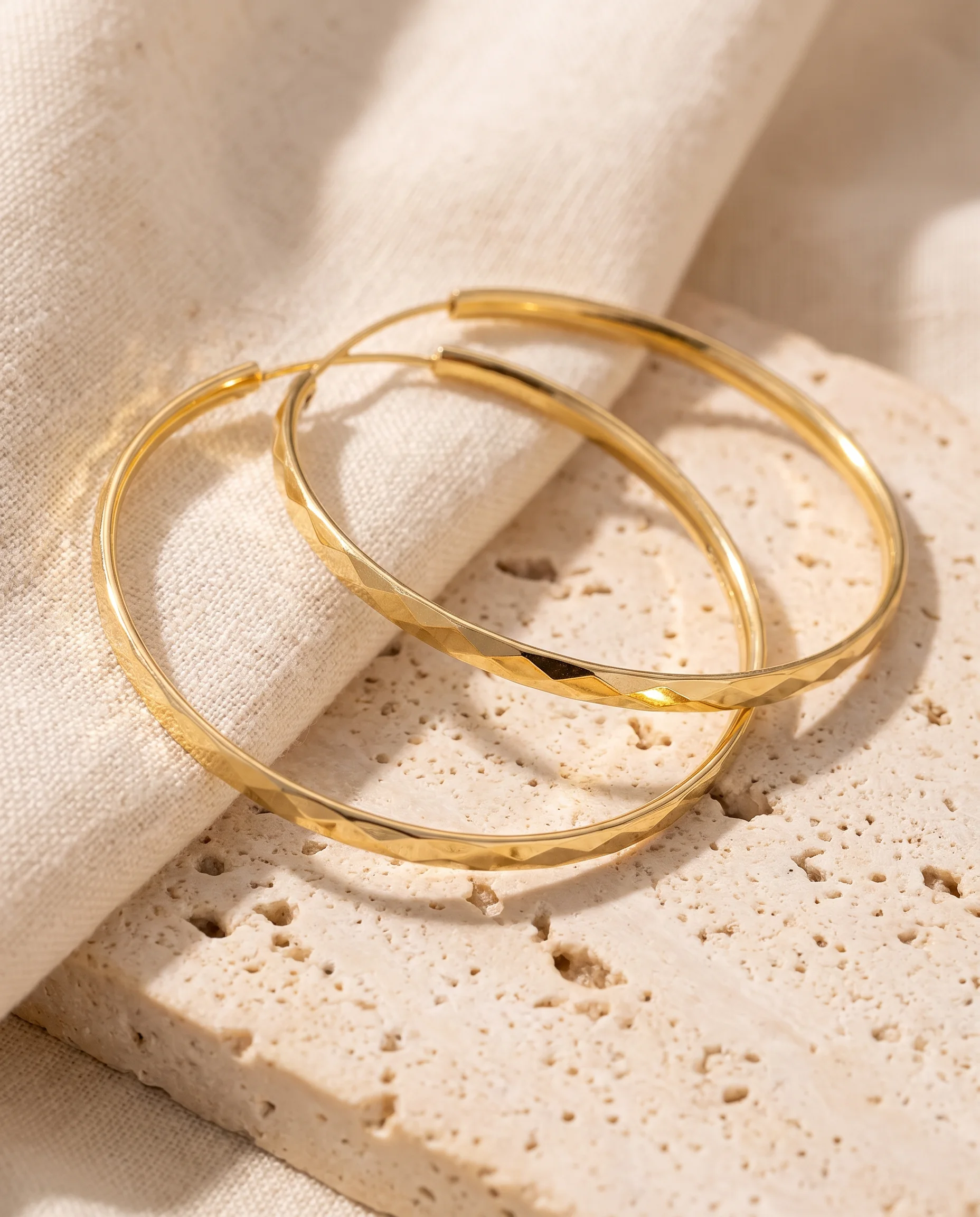 Large gold faceted hoops placed on a light stone, near ecru linen