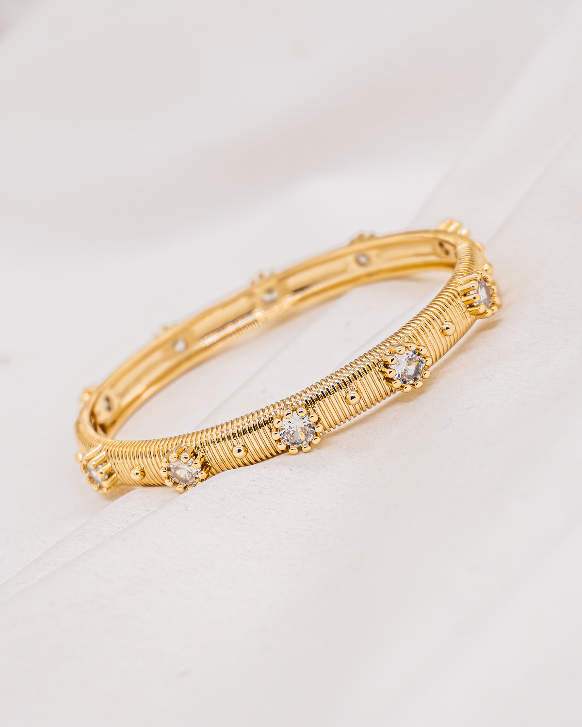 Bracelete dourado estriado com finas listras, cravejado com pedras claras, sobre um tecido off-white