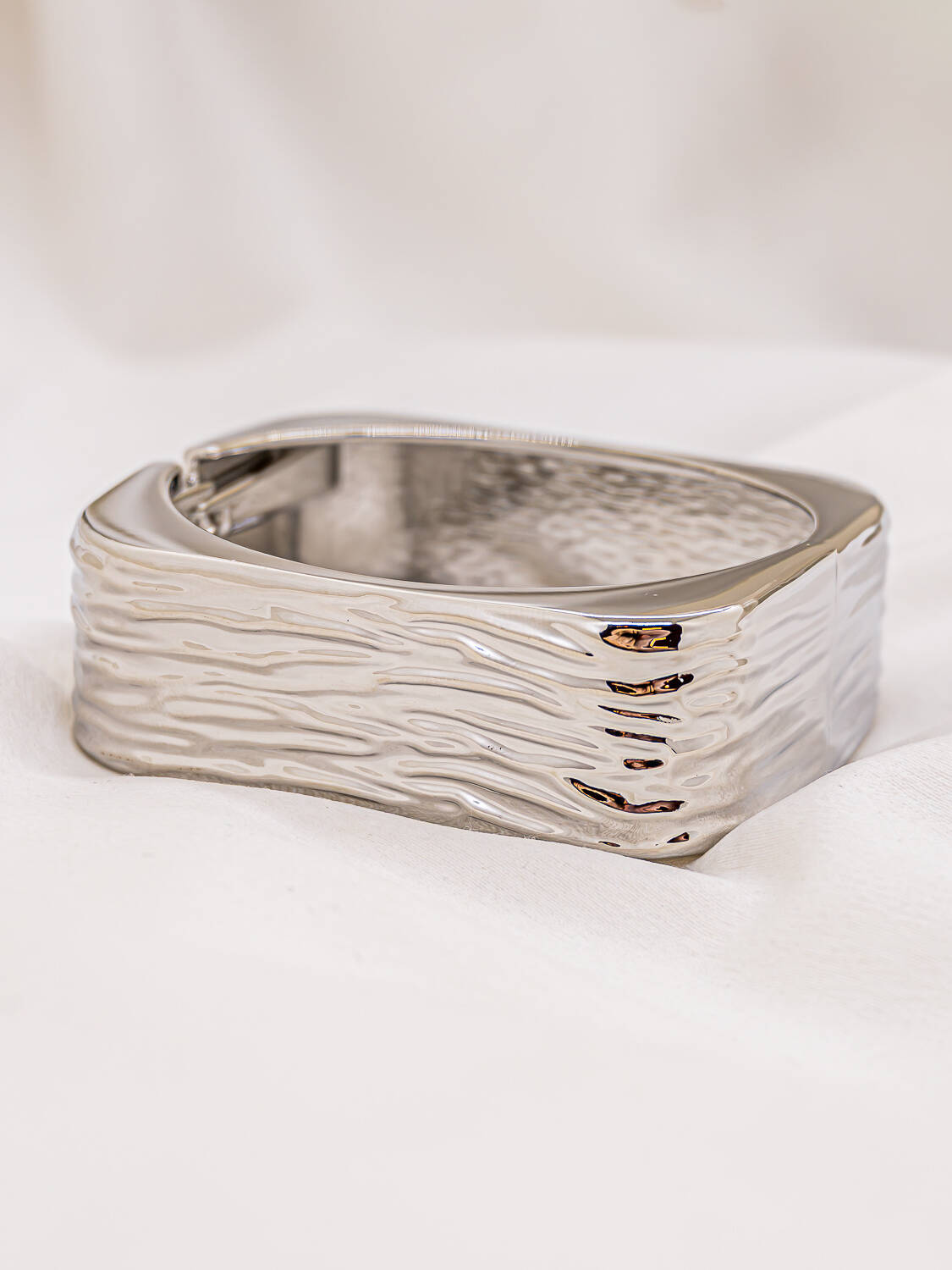 Large jonc argenté de forme carrée à surface ondulée et brillante, posé sur tissu blanc